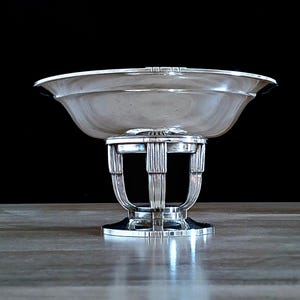 May include: A silver-toned, Art Deco-style bowl on a pedestal. The bowl has a wide, shallow design with a flared rim. The pedestal features vertical bars and a circular base. The background is black.