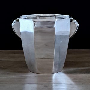 Op de afbeelding: Een zilveren ijsemmer met een geometrisch ontwerp. De emmer heeft een facetgeslepen body en twee gebogen handvatten. De binnenkant is niet zichtbaar. De achtergrond is zwart en de emmer staat op een houten oppervlak.