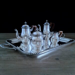 Könnte beinhalten: Ein silbernes Teeservice mit Tablett. Das Set enthält eine Teekanne, einen Sahnekännchen, eine Zuckerdose und zwei weitere Behälter. Die Teekanne und das Sahnekännchen haben Holzgriffe. Das Tablett hat einen Perlenrand und Griffe.