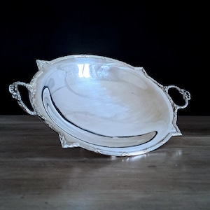 May include: A silver-colored, oval serving dish with ornate handles and a decorative rim. The dish has a reflective surface and is set against a dark background. The handles have a detailed, floral design. The dish is likely made of metal.
