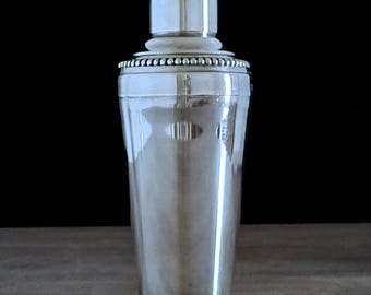 Puede incluir: Un coctelera de plata con un borde de cuentas y una parte superior blanca. La coctelera está sobre una superficie de madera.