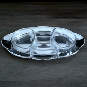 May include: A clear glass serving dish with multiple compartments, set on a silver-colored metal base. The dish has an oval shape with rounded edges and is designed for serving appetizers or snacks. The compartments are various sizes.