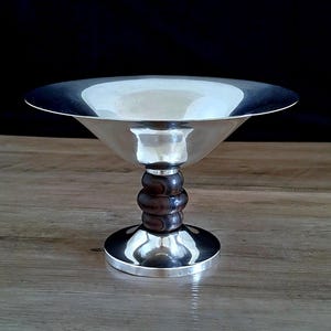 May include: A silver bowl with a wide, shallow base and a wooden stem. The bowl is polished and reflects light. The stem is made of dark wood with a striped pattern.