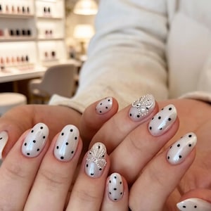 Pode incluir: Unhas ovais brancas peroladas com bolinhas pretas. Uma unha apresenta um design de laço prateado com pequenas pérolas. As unhas são bem cuidadas e têm um acabamento brilhante, mostrando um estilo de nail art clássico e elegante.