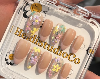 May include: A set of nude-colored press-on nails with floral designs in shades of pink, yellow, and green. The nails are arranged in a clear plastic case with the text "HeidiStudioCo" printed on the front.