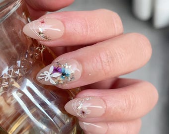 May include: Close-up of a hand with almond-shaped nails painted a light nude color. Each nail features a delicate floral design with dried flowers and glitter accents.