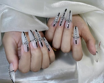 Pode incluir: Close-up de unhas longas e em forma de amêndoa, brancas, com linhas pretas e strass formando um design de cruz.