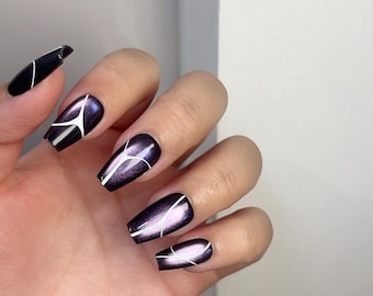 Op de afbeelding: Een hand met lange, doodskistvormige kunstnagels. De nagels zijn diep paars met een metallic glans en een wit geometrisch ontwerp. Het ontwerp bestaat uit dunne, hoekige lijnen die een modern, abstract patroon creëren.
