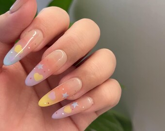 Puede incluir: Una mano con uñas acrílicas largas en forma de almendra. Las uñas presentan un diseño degradado con tonos amarillos, naranjas y celeste. Cada uña está decorada con pequeños corazones amarillos y estrellas azules.