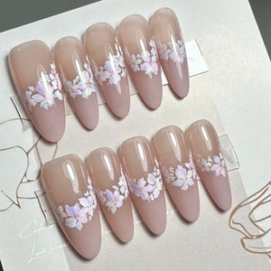 May include: Set of ten nude-colored false nails with a white floral design. The nails are long and almond shaped.