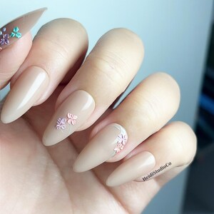 May include: Almond-shaped, beige artificial nails. Some nails feature small, three-dimensional floral designs in pastel colours. The text "HeidiStudioCo" is visible on one nail.