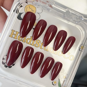Puede incluir: Un juego de diez uñas postizas ovaladas de color burdeos en una caja de plástico transparente. Las uñas son brillantes y tienen un acabado liso. La caja tiene un diseño floral y el texto "Heidi Studio Co."