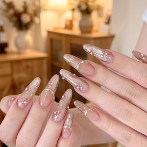 May include: Long, almond-shaped artificial nails with a nude base and intricate floral designs in pink and gold. Some nails feature a silver beaded accent at the base. The nails are a light, translucent color.