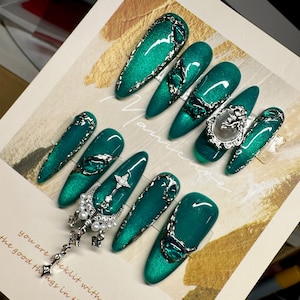 May include: Set of ten emerald green press-on nails with silver accents and embellishments. Some nails feature a silver crescent moon, a star, and a key charm. Other nails have a silver filigree design and a green gem. One nail has a silver frame with a white cameo.