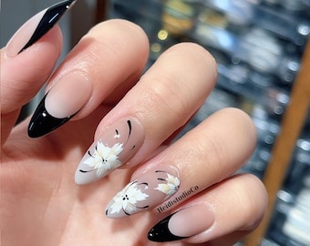 Könnte beinhalten: Mandelförmige Acrylnägel mit schwarzem und nudefarbenem French-Tip-Design. Einige Nägel weisen weiße Blumenmuster mit schwarzen Akzenten auf. Der Text "HeidistudioCo" ist auf einem Nagel sichtbar.