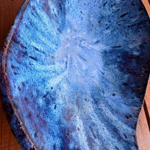 May include: A handmade ceramic serving dish with an organic shape. The dish has a striking blue glaze, with variations from deep indigo to light sky blue. The surface has a textured appearance with subtle speckles. A unique piece.