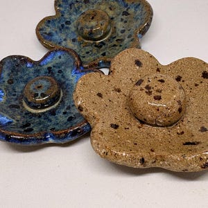 May include: Three ceramic flower-shaped incense holders. Two are blue with dark speckles and a glossy finish, and one is tan with brown speckles. Each has a central raised area with a hole for incense.