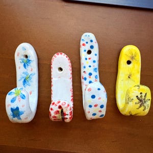 May include: Four ceramic wall hooks. The hooks are white, red, and yellow, with painted floral and polka dot designs. Each hook has a hole for mounting. The hooks are on a brown surface.