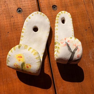 May include: Two handmade ceramic wall hooks. One hook features a yellow flower design, the other a tree with pink blossoms. Both are white with green and yellow accents and a hole for hanging.