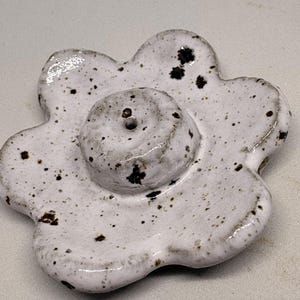 May include: A white ceramic flower-shaped object with a central, raised circular element. The piece has a speckled pattern of dark brown spots and a glossy finish. The flower has six rounded petals.