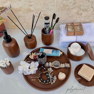 May include: A selection of brown bathroom accessories, including a soap dispenser, reed diffuser, toothbrush holder, and a tray with a watch, jewellery, and a seashell. Other items include cotton pads, soap, and a hairbrush.