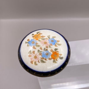Puede incluir: Un botón de porcelana blanca con un diseño floral azul y dorado. El botón tiene un borde azul oscuro y está decorado con un grupo de flores en tonos azules, rosas y naranjas.