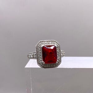 May include: A silver ring with a large, red, rectangular gemstone surrounded by smaller, clear gemstones.