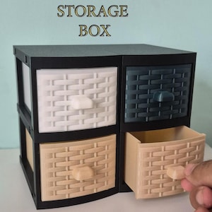 Pode incluir: Uma caixa de armazenamento interligada com quatro gavetas em branco, bege e azul escuro, com uma moldura preta. O texto "INTERLOCKING STORAGE BOX" está no topo. Uma gaveta está aberta.