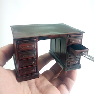 May include: A small, dark brown wooden desk with a classic design. It features multiple drawers, with one drawer open. The desk is held in a hand. The desk has a rectangular top and a series of drawers on both sides.