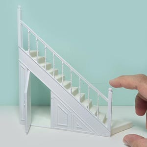 Puede incluir: Una escalera en miniatura blanca con una pequeña puerta que se abre en la parte inferior. La escalera tiene una barandilla blanca y está colocada sobre un fondo azul claro.