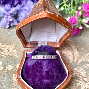 May include: A vintage ring with alternating red and clear stones, displayed in a brown leatherette jewelry box with a purple velvet interior. The box is open, revealing the ring. The background features purple and pink flowers.