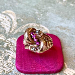 Peut inclure: Une bague dorée au design tourbillonnant, ornée de trois petites pierres précieuses roses vives. La bague est présentée sur une surface de velours rose vif, mettant en valeur ses détails complexes et sa finition polie.