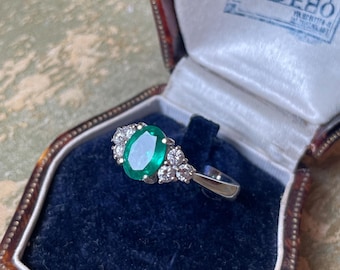 May include: An oval emerald ring with a halo of small diamonds, set in a silver band, rests in a vintage jewelry box. The ring's emerald is a vibrant green, and the diamonds sparkle. The jewelry box is lined with blue velvet.