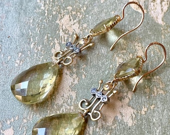 May include: A pair of gold-toned drop earrings with pear-shaped, faceted, pale yellow gemstones. The earrings feature intricate filigree and small, square, silver-toned accents. The ear wires are simple gold hooks.