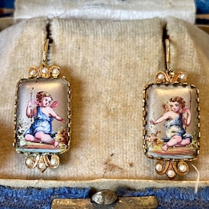 May include: A pair of gold earrings with a rectangular enamel design featuring a cherub in a blue dress. The earrings are accented with small pearls.