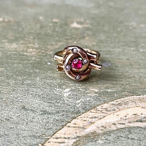 Puede incluir: Un anillo dorado con una gema roja central y pequeñas perlas blancas. El anillo tiene un diseño intrincado y arremolinado, con una estética vintage. El anillo es una joya.