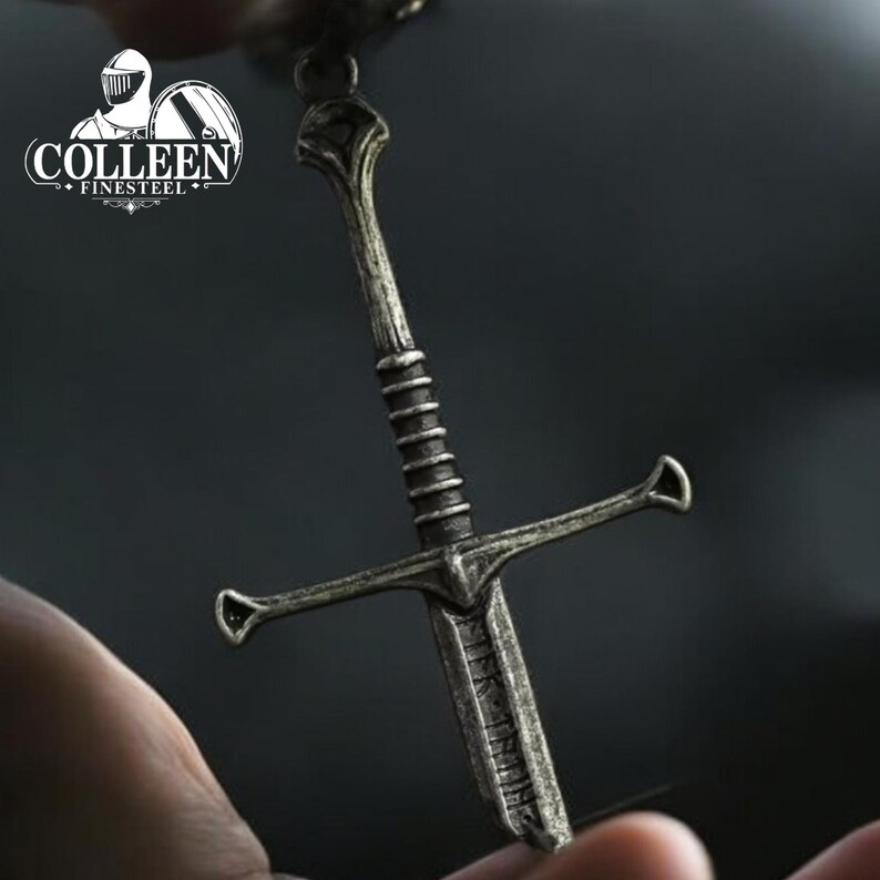 May include: A silver-toned sword pendant with a detailed hilt and blade. The pendant is hanging from a chain.