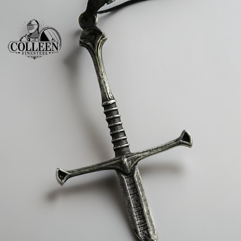 May include: A silver-toned sword pendant with a detailed design. The sword has a cross guard and a pointed tip. The pendant is attached to a black cord.