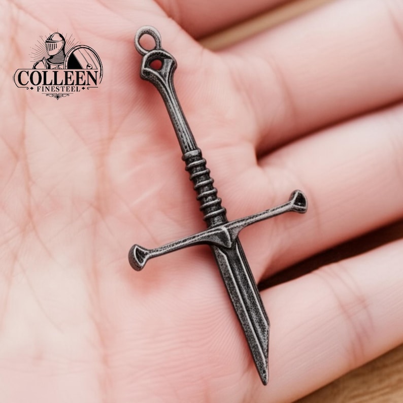 May include: A silver-toned sword pendant with a detailed design. The sword has a cross guard and a pointed tip. The pendant is on a hand.