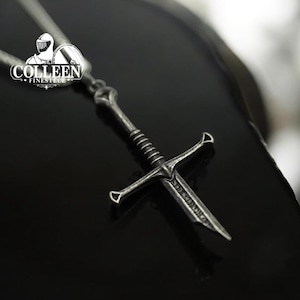 May include: A silver sword pendant necklace with a detailed design. The pendant is shaped like a sword with a cross guard and a pointed tip. The necklace is on a black background.