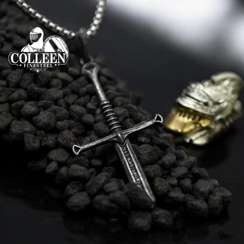 May include: A silver sword pendant necklace with a detailed design. The sword is hanging on a chain and is resting on a bed of black stones. The pendant is a replica of a medieval sword.