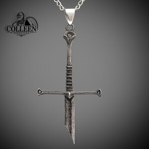May include: A silver sword pendant necklace with a detailed design. The sword has a cross guard and a pointed tip. The necklace is on a silver chain.