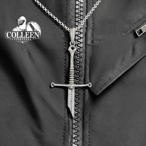 May include: A silver sword pendant necklace on a silver chain. The sword has a cross guard and a pointed tip. The pendant is hanging from a zipper on a black jacket.