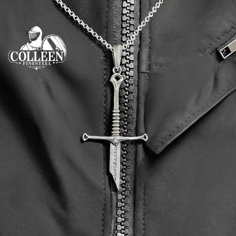 May include: A silver sword pendant necklace on a silver chain. The sword has a cross guard and a pointed tip. The pendant is hanging from a zipper on a black jacket.