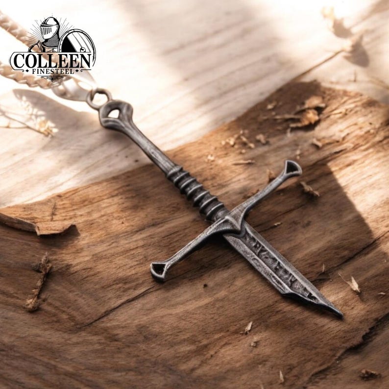 May include: A silver sword pendant necklace with a detailed design. The pendant is on a wooden surface with the text "COLLEEN FINESTEEL" on the necklace.
