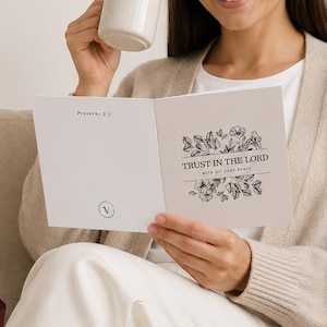 May include: A white greeting card featuring a black floral illustration and the text "TRUST IN THE LORD WITH ALL YOUR HEART" and "Proverbs 3:5". The card is held open, displaying a small circular logo with the letter V on the bottom left corner.
