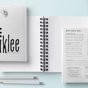 May include: White spiral-bound journal with the word "Wiklee" in bold black letters. The journal includes a brain graphic and a weekly check-in page. Two pencils are placed below the journal.
