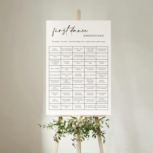 Può includere: Un cartello bianco su un cavalletto di legno con la scritta "first dance" in elegante corsivo. Il cartello è un concorso a premi con titoli di canzoni e spazio per scrivere i nomi. Il cavalletto è decorato con verde.