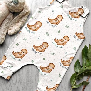 K&ouml;nnte beinhalten: Wei&szlig;er Baby-Strampler mit einem Muster aus braunen Ottern, die Fische halten, gr&uuml;nen Bl&auml;ttern und blauen Sternen. Der Strampler hat den Text "Otterly Cute" wiederholt &uuml;ber das gesamte Muster.