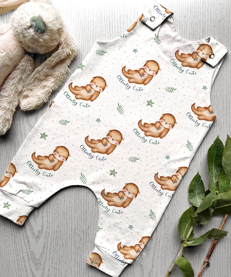 K&ouml;nnte beinhalten: Wei&szlig;er Baby-Strampler mit einem Muster aus braunen Ottern, die Fische halten, gr&uuml;nen Bl&auml;ttern und blauen Sternen. Der Strampler hat den Text "Otterly Cute" wiederholt &uuml;ber das gesamte Muster.